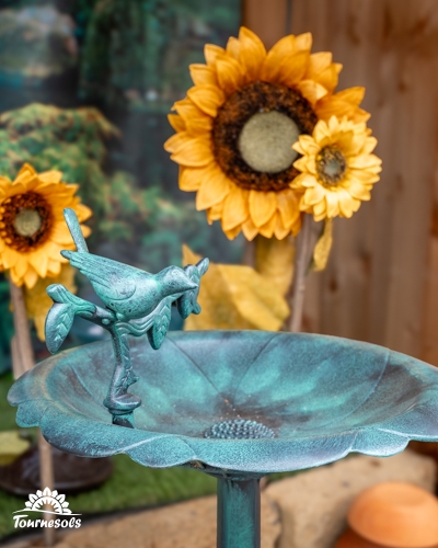 Bain d'oiseaux en fonte vert-de-gris orné d'un oiseau décoratif, exposé devant des tournesols artificiels en jardinerie